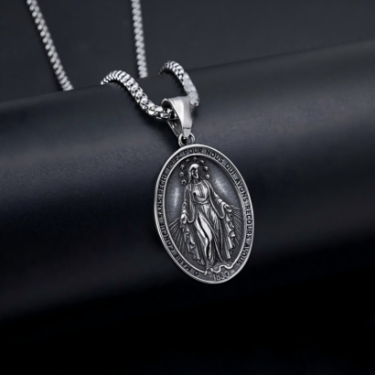 A studio product shot of a silver-toned stainless steel oval Miraculous Medal pendant necklace resting on a dark surface, featuring an intricate relief of the Virgin Mary surrounded by French inscription text, the year 1830, and a matching chain.