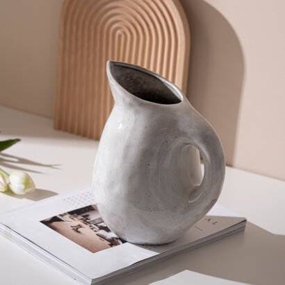 Off-white cream ceramic pitcher vase with organic shape and integrated curved handle resting on a magazine