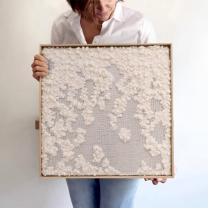 A square wooden-framed textile wall art piece held by a person, featuring organic cloud-like clusters of textured white wool woven and layered across a semi-transparent grid mesh background.