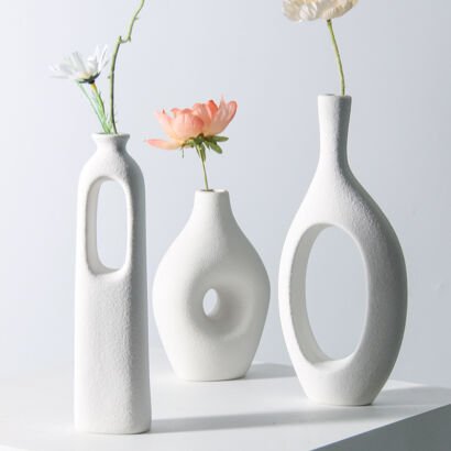 Three matte white hollow ceramic vases with geometric cutouts displaying single delicate flower stems on a white table