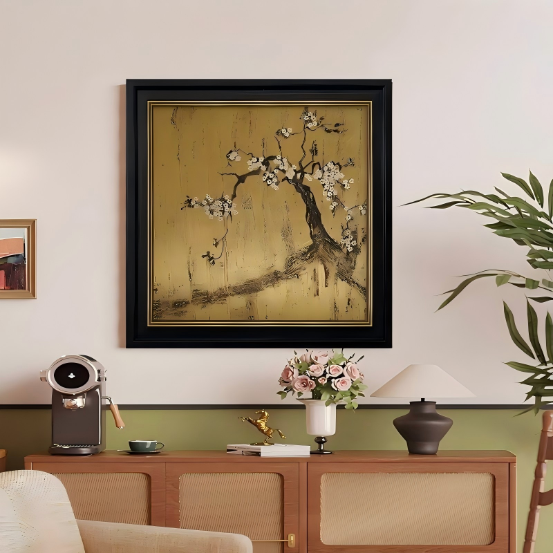 A square framed art piece hanging on a warm neutral wall above a wooden media console, featuring a dark blossoming plum tree branch sprawling across a textured metallic gold leaf background inside a black and gold frame.