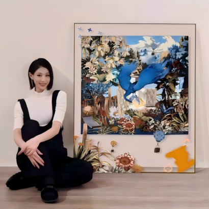 A large slim-framed 3D collage wall art artwork resting on a wooden floor alongside a seated woman, featuring a central surreal artwork with a vibrant blue bird in mid-flight surrounded by lush botanical elements, hot air balloons, and floating dimensional cutouts extending past the canvas border.