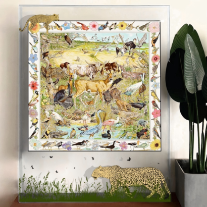 A large transparent acrylic display frame set against a wall next to a potted green plant, featuring a central silk scarf illustration of Texas wildlife including deer, longhorns, and birds, decorated with floating leopard graphics and grass silhouettes on the clear outer border.