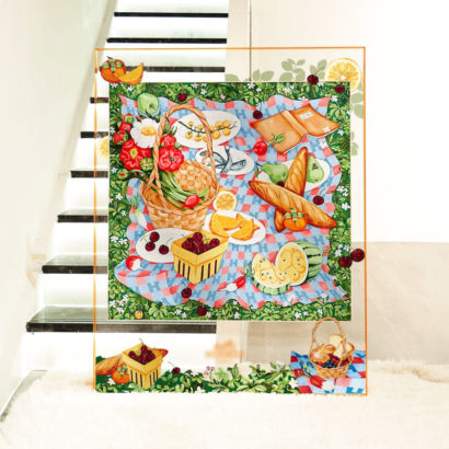 A large transparent acrylic art display frame resting on a plush white rug near a staircase, featuring a central silk scarf illustration of a vibrant summer picnic spread with fruits, bread baguettes, and woven baskets on a blue checkered blanket, with floating fruit and basket motifs extended onto the outer clear frame border.