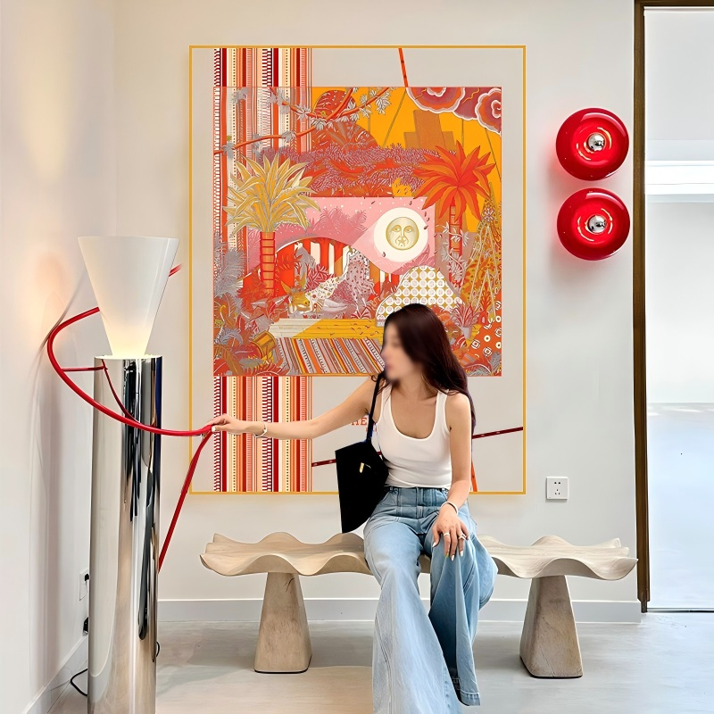 A large transparent acrylic art display frame mounted on a modern white wall above a wooden bench, featuring a central silk scarf illustration in warm orange, red, and yellow tones depicting palms, desert landscapes, and a radiant sun face motif, with a red sculptural line graphic extending beyond the frame.