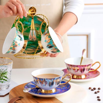 Colorful ceramic teacups with abstract brushstroke pattern and gold rim sitting on matching saucers alongside a metal display rack holding additional cups and spoons