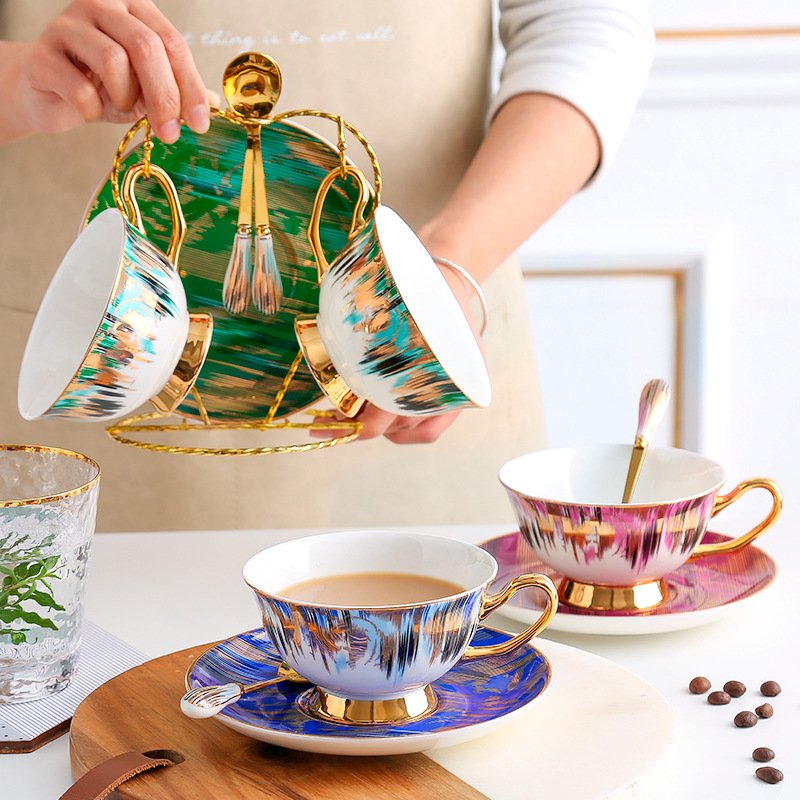 Colorful ceramic teacups with abstract brushstroke pattern and gold rim sitting on matching saucers alongside a metal display rack holding additional cups and spoons