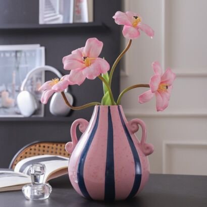 A rounded pink ceramic vase with vertical dark blue stripes, textured speckling, and elegant side handles resting on a dark tabletop, filled with an arrangement of light pink open-petaled flowers, with an open book and a small glass candle holder nearby.