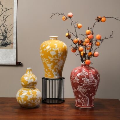 A collection of three ceramic vases featuring white floral patterns against yellow and red backgrounds, including a gourd-shaped yellow vase, an elevated yellow vase on a black metal stand, and a red vase filled with decorative branches with small orange fruits, displayed on a wooden table against a neutral wall with a hanging scroll.