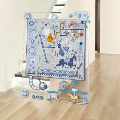 A large transparent acrylic art display standing on a light wooden floor near a hallway, featuring a framed silk scarf illustration of a whimsical blue mechanical circus machine with an astronaut, horse, and floating balloon accents on the outer frame.
