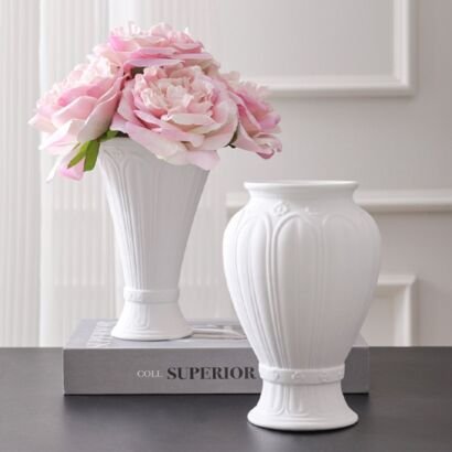 Two white ceramic vases with ribbed and embossed botanical details displayed together on a table, with the taller vase placed on top of a book and filled with an arrangement of light pink peonies.