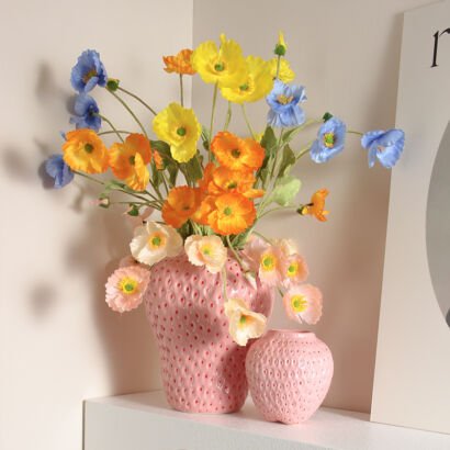 Two pink ceramic vases shaped like strawberries with textured seed details displayed on a white shelf, with the larger vase filled with a vibrant mixed arrangement of colorful blue, yellow, orange, and pale pink poppies.
