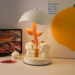 Charming desk lamp with an umbrella-shaped lampshade over an orange plant stem, featuring two white cartoon elephants wearing golden crowns resting at the base