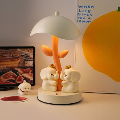 Charming desk lamp with an umbrella-shaped lampshade over an orange plant stem, featuring two white cartoon elephants wearing golden crowns resting at the base