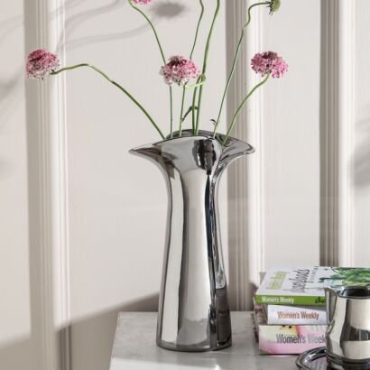 A sleek, shiny silver metallic vase with a unique flared rim standing upright on a white tabletop against a white paneled wall, holding long-stemmed pink flowers, with stacked magazines beside it.