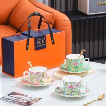 Three ceramic teacups with matching saucers and golden spoons displayed on a table alongside an orange and blue gift box branded with "CHAO HAO fine bone china", featuring delicate floral and bird patterns in pink, green, and blue.