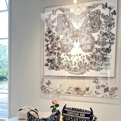 A large square acrylic floating frame displaying a fine silk scarf with monochrome botanical and butterfly artwork, mounted on a neutral wall above designer handbags.