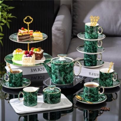 An elaborate luxury porcelain tea set in a rich malachite green marble pattern, featuring a teapot, sugar bowl, creamer, matching teacups with saucers and gold spoons, a stacked tier of cups, and a two-tier dessert stand holding various small cakes.