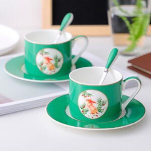 Two vibrant emerald green ceramic teacups with matching saucers and spoons sitting on a white surface, featuring a circular medallion on the front displaying a cute illustration of a little bunny surrounded by green foliage and red berries.