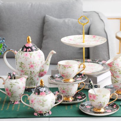 A complete vintage porcelain tea set arranged on a tabletop, featuring a teapot, sugar bowl, creamer, teacups with saucers, spoons, and a two-tier serving stand, all adorned with pink and purple floral patterns against a white and black background.