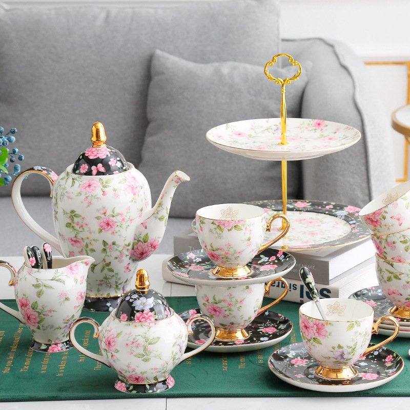 A complete vintage porcelain tea set arranged on a tabletop, featuring a teapot, sugar bowl, creamer, teacups with saucers, spoons, and a two-tier serving stand, all adorned with pink and purple floral patterns against a white and black background.