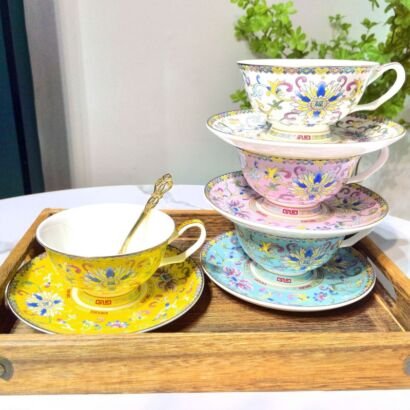 A collection of vibrant ceramic teacups with matching saucers and gold spoons displayed on a wooden tray and a stacked tier, featuring intricate colorful floral and geometric patterns inspired by traditional Chinese cloisonné enamel art.