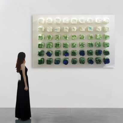 A large white canvas with a grid array of 3D textured cubes transitioning in color from cream white to forest green and deep blue accents, displayed on a wall in a modern gallery setting with a woman viewing it.