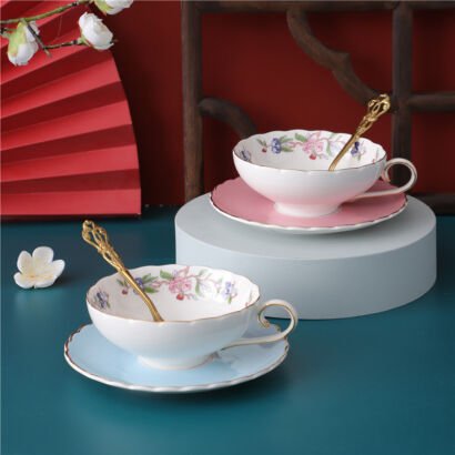 Two elegant ceramic teacups with matching saucers and ornate gold spoons arranged on a tabletop, featuring scalloped rims, delicate floral garlands inside the cups, and pastel pink and blue bases.