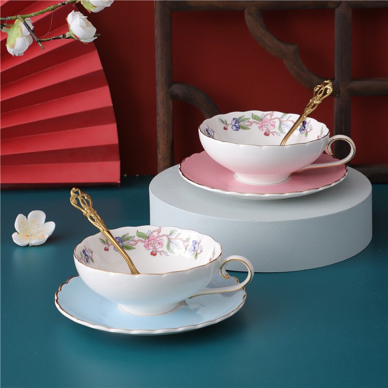 Two elegant ceramic teacups with matching saucers and ornate gold spoons arranged on a tabletop, featuring scalloped rims, delicate floral garlands inside the cups, and pastel pink and blue bases.