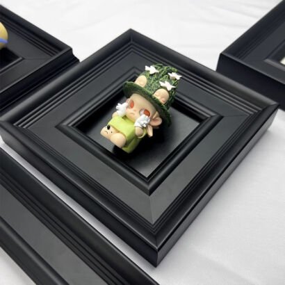 A square black stepped picture frame mounted on a white surface, containing a cute stylized designer art toy figure of a green elf wearing a textured green hat adorned with tiny white flower details.
