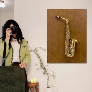 A vertical textured canvas wall art hanging on a neutral wall next to a person holding a camera, featuring a realistic 3D golden brass saxophone relief mounted against a warm brown distressed background.