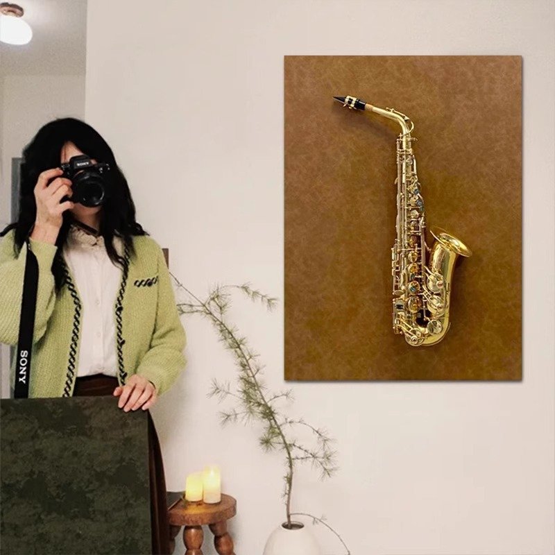 A vertical textured canvas wall art hanging on a neutral wall next to a person holding a camera, featuring a realistic 3D golden brass saxophone relief mounted against a warm brown distressed background.