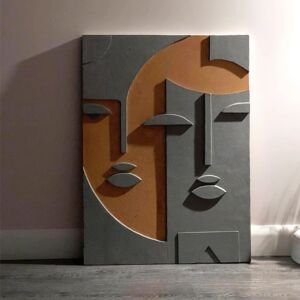 A vertical abstract 3D relief wall sculpture resting on the floor against a neutral wall, featuring a stylized cubist composition of overlapping human faces rendered in textured dark grey plaster and warm terracotta brown tones.