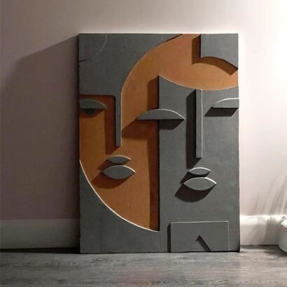 A vertical abstract 3D relief wall sculpture resting on the floor against a neutral wall, featuring a stylized cubist composition of overlapping human faces rendered in textured dark grey plaster and warm terracotta brown tones.