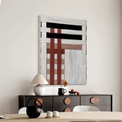A vertical textured abstract wall art piece hanging on a plain neutral wall above a dark sideboard cabinet, featuring an intricate dimensional geometric design made from layered white, black, and terracotta red threads and fibers.