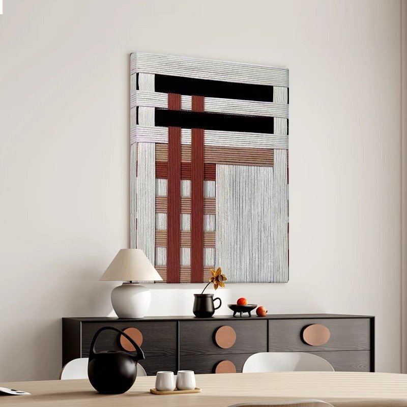 A vertical textured abstract wall art piece hanging on a plain neutral wall above a dark sideboard cabinet, featuring an intricate dimensional geometric design made from layered white, black, and terracotta red threads and fibers.