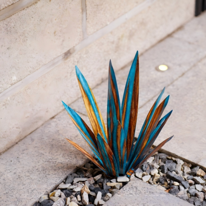 Metal agave yard art sculpture, blue and rust gradient painted metal agave plant decor placed in gravel stone paving gap
