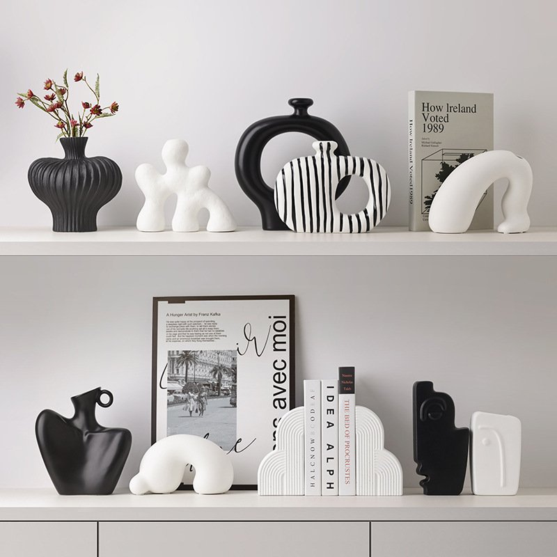Collection of modern abstract black and white ceramic vases, hollow donut vessels, arch sculptures, and bookends arranged on minimalist white display shelves