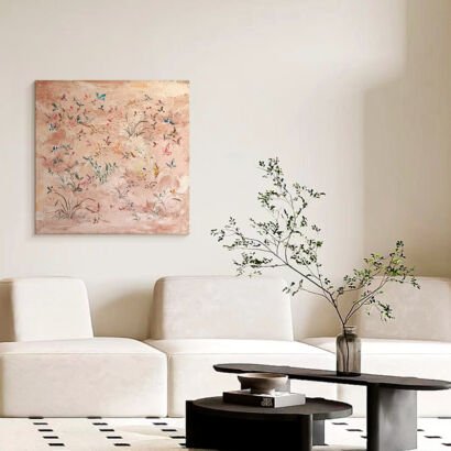 A square artwork with a textured blush pink background depicting delicate wildflowers and colorful butterflies, mounted on a white wall above a modern white modular sofa and a low coffee table with a vase.