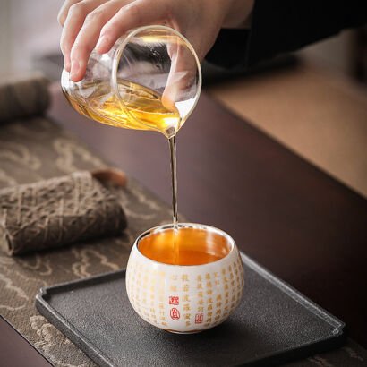 A clear glass pitcher pouring warm golden tea into a white ceramic teacup with a silver-lined interior, featuring gold Chinese Heart Sutra calligraphy and red seal stamps, resting on a dark slate tray.