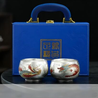 Two handcrafted silver teacups featuring vibrant dragon and phoenix enamel motifs, set in front of a blue suitcase-style luxury gift box with a top carrying handle and gold seal emblem.
