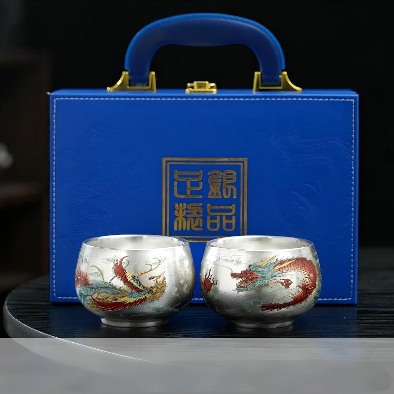 Two handcrafted silver teacups featuring vibrant dragon and phoenix enamel motifs, set in front of a blue suitcase-style luxury gift box with a top carrying handle and gold seal emblem.
