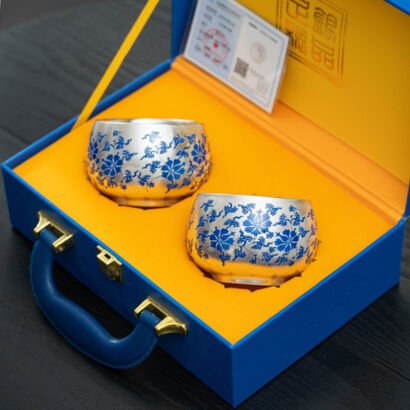 Two polished silver teacups adorned with blue enamel floral scrollwork, presented inside a vibrant blue and yellow luxury suitcase-style gift box with a leather handle and official certificate card.