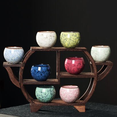Eight small rounded ceramic teacups with colorful kiln-transformed crackle glazes displayed on a dark wooden teapot-shaped curio shelf against a black background, featuring shades of sky blue, white, yellow-green, mint, cobalt, crimson, jade, and pink.
