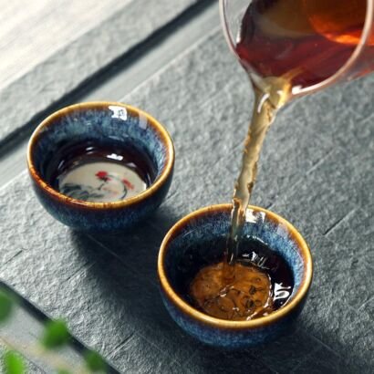 Amber tea pouring from a glass pitcher into one of two small handcrafted ceramic teacups resting on a dark stone tea tray, highlighting deep ocean blue speckled glazes, golden-brown rims, and a handpainted motif inside the bottom.