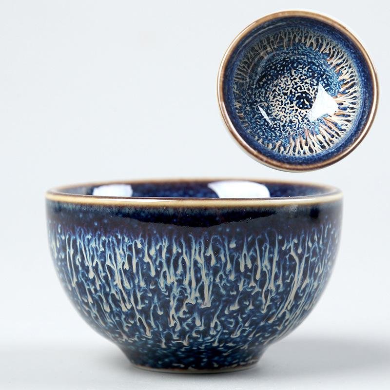 A side view and top-down view of a handcrafted small ceramic teacup with a deep midnight blue glaze, intricate cream lightning-like drip textures, and a golden-brown rim set against a plain background.