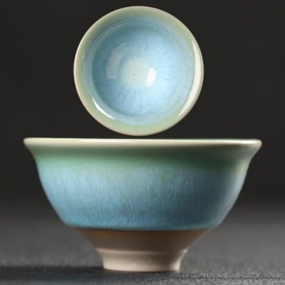 A side view and top-down view of a handcrafted ceramic teacup with a gradient sky-blue to celadon green glaze, fine radial streak textures, and an unglazed pale foot set against a dark background.