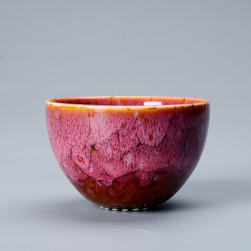 A side view of a rounded ceramic Jian Zhan teacup set against a neutral grey background, showcasing a vibrant magenta pink reactive glaze with textured drips over a deep amber base and a golden rim.