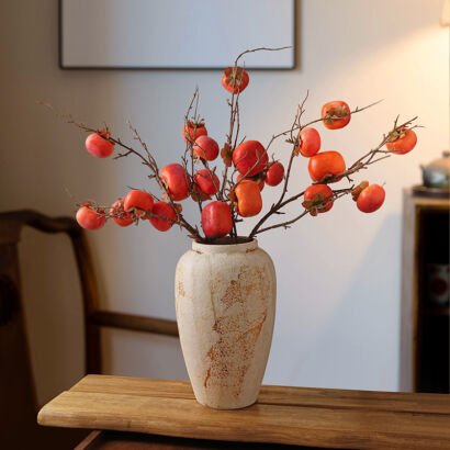 Faux persimmon branches laden with bright orange fruit arranged in a rustic distressed beige ceramic vase on a wooden bench