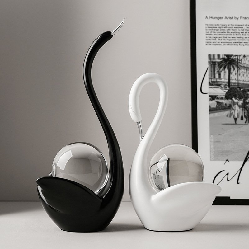 Pair of sleek black and white swan sculptures with metallic silver beaks cradling clear crystal spheres on a tabletop next to framed wall art